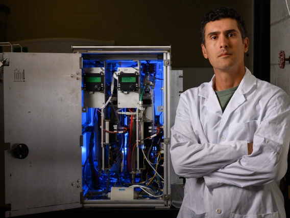 A man in a white lab coat stand in front of a blue-lit machine.