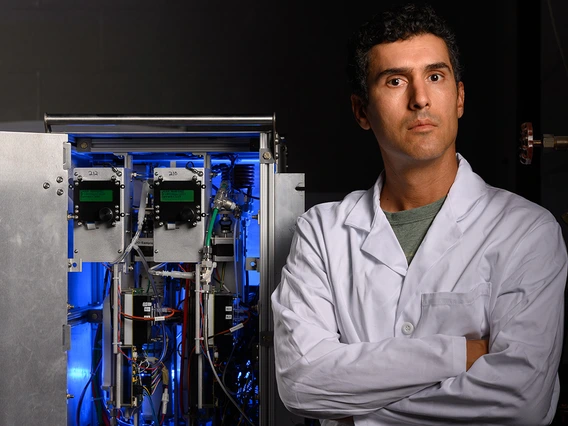 A man with dark hair in a lab photo stands i front of a blue machine.