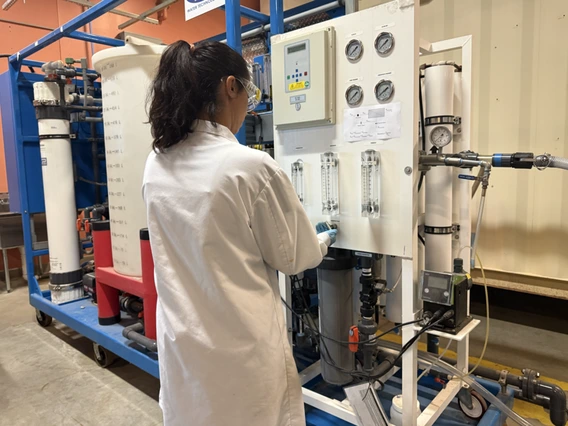 Researcher in white lab coat stands in front of water treatment equipment