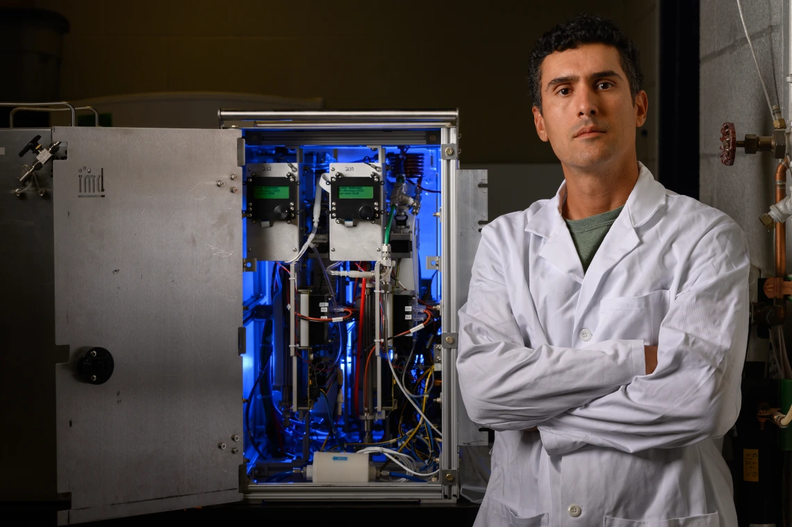 A man in a white lab coat stand in front of a blue-lit machine.